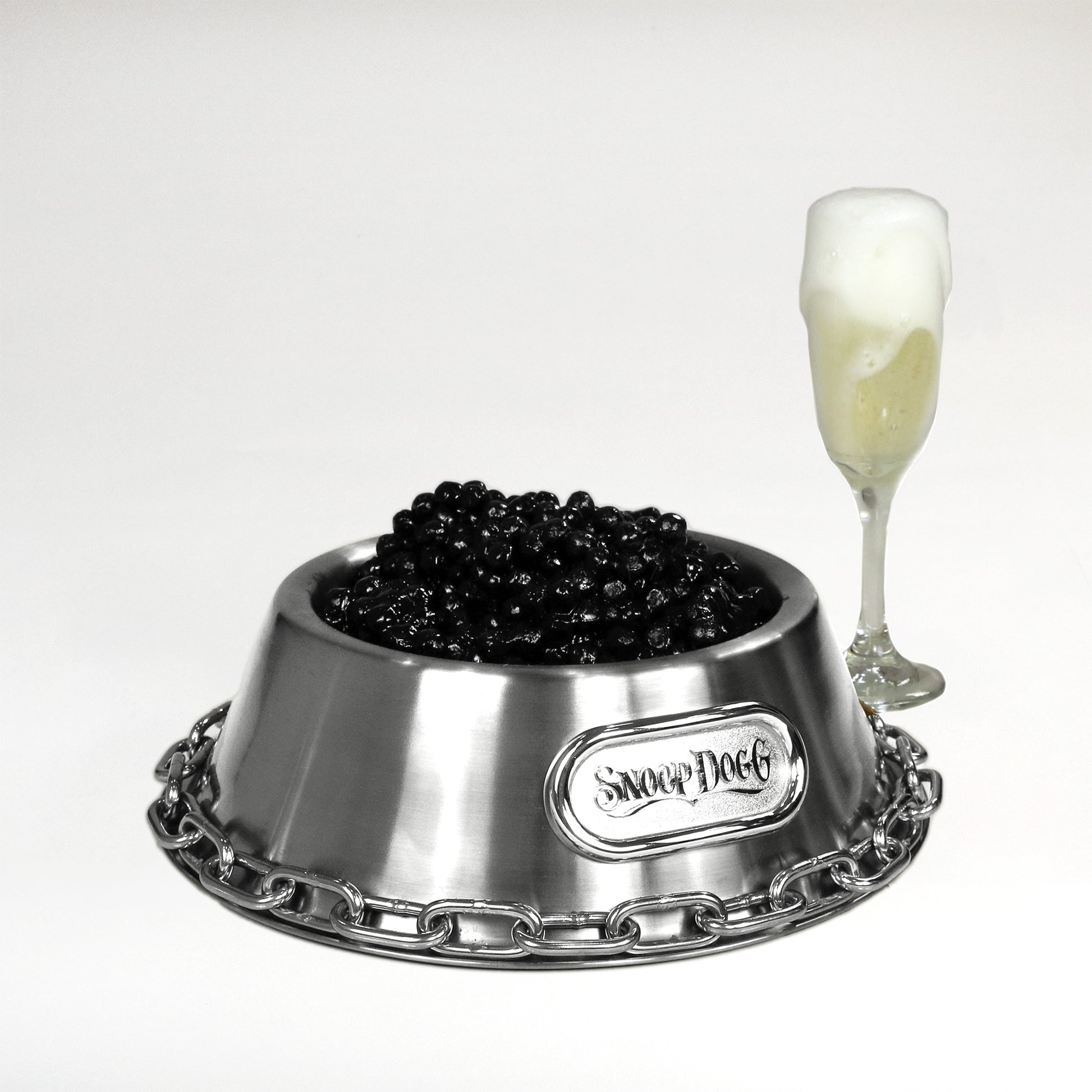 Product flat lay of the Large Deluxe Silver Pet Bowl filled with caviar sitting next to a glass of champagne.